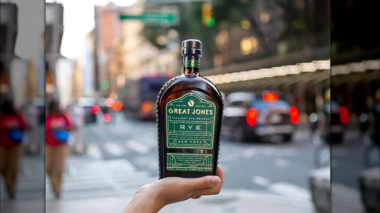 Hand holding a bottle of Great Jones Rye outdoors with a city streets in the background