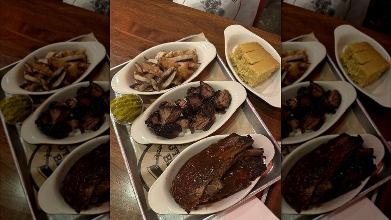 Pork belly, burnt ends, and ribs on small plates on a metal tray, with a small cup of pickles and a dish of cornbread.