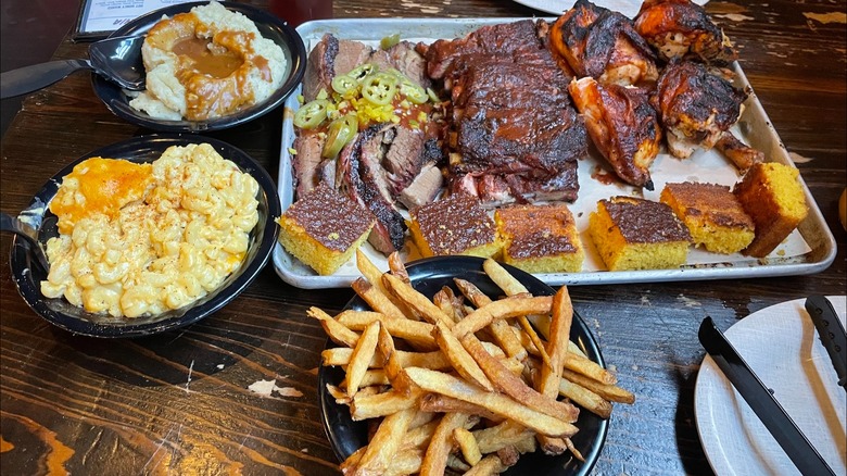 A large tray of BBQ, featuring brisket, ribs, chicken, and cornbread, with sides like mac and cheese, mashed potatoes, and French fries on a scratched wooden table.