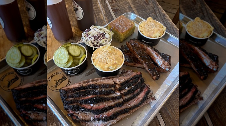 Sliced BBQ brisket and ribs on a tray with various sides, including pickles, mac and cheese, cornbread, and coleslaw.