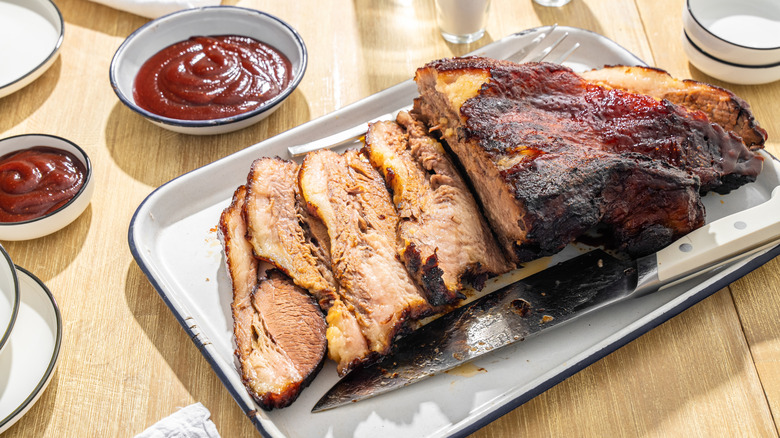 Partially sliced barbecue brisket on a metal tray, accompanied by small cups of sauce.