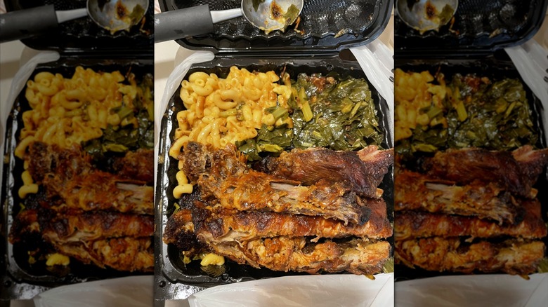 Pork ribs, creamy mac and cheese, and collard greens piled high in a black plastic takeout container.