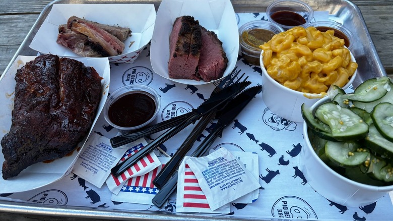 A tray of BBQ meats, plus mac and cheese, pickles, and an assortment of sauces, on a wooden table.