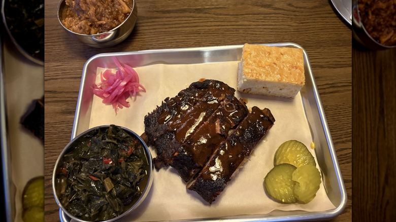 Ribs with BBQ sauce, collard greens, cornbread, pickles, and pickled onions sit atop a piece of brown paper on a metal tray.