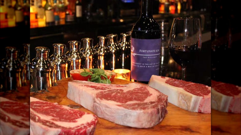 Raw steaks on wooden board in front of wine bottle, glass, and cocktail shakers