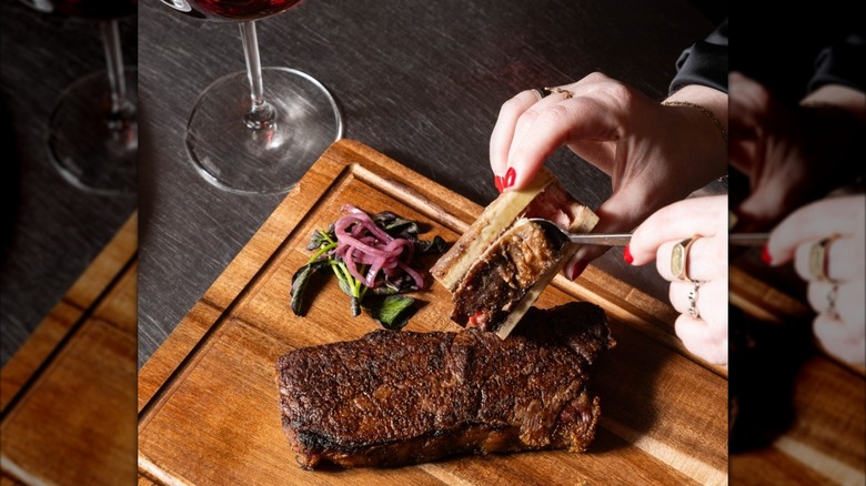 Scooping bone marrow onto steak on a wooden board with glass of wine