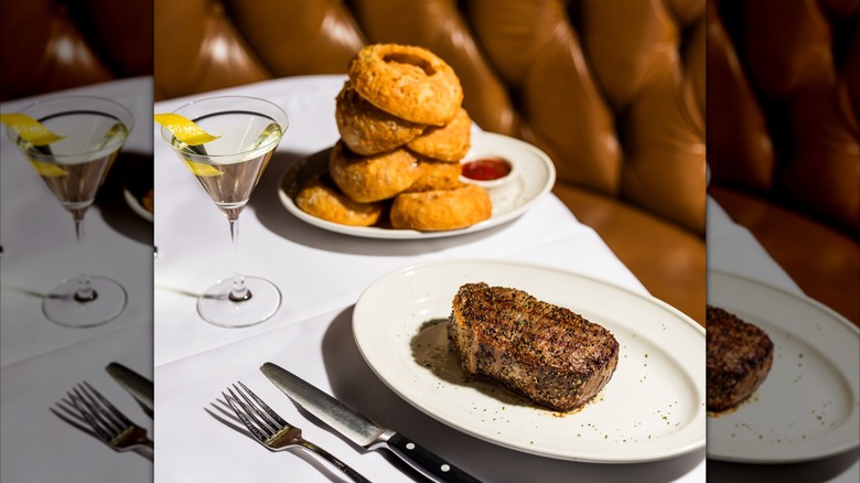 Cocktail and plates with steak and onion rings on white tablecloth