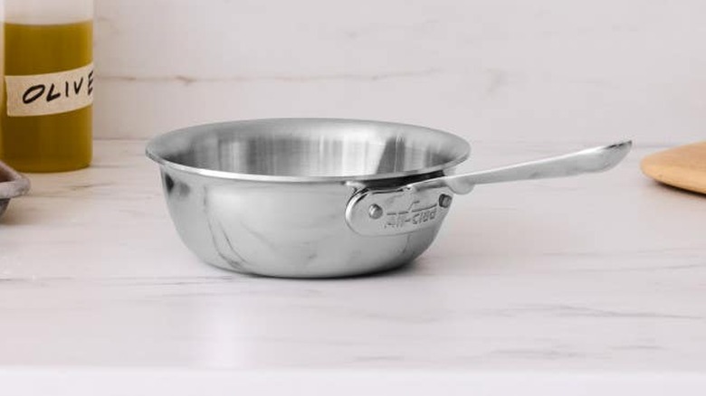 An All-Clad saucier on a marble counter with olive oil seen in the background