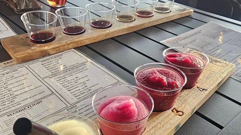 A bright-colored wine slushie flight in the foreground with a wine flight in the background, both served in clear cups on wooden boards,  on a table with wine menus.