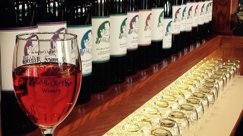 A single line of bottles of Bear Creek Winery wines on a bar shelf with a winery-branded glass of light colored wine in the foreground, and rows of stemless glasses on white matting on a shelf below.
