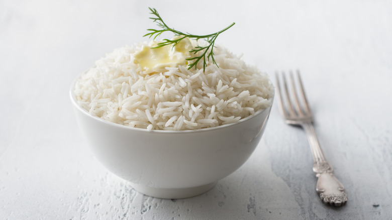 Small white bowl of cooked long grain white rice topped with butter and green garnish next to silver fork