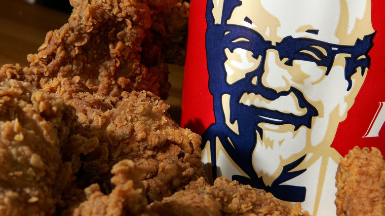 A pile of KFC fried chicken stacked in front of Colonel Sanders' face on the side of a KFC bucket.