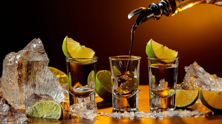 Liquor pouring from a bottle into a row of shot glasses, two of which are topped with lime slices, which chunks of ice around them
