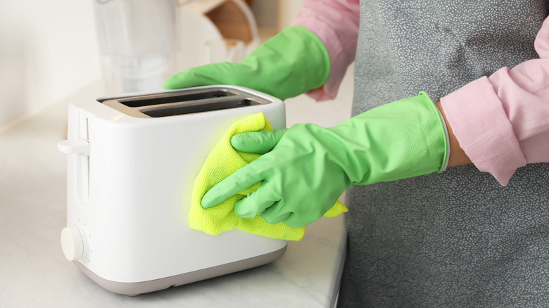 A woman wearing rubber gloves and an apron cleaning a toaster with a damp cloth