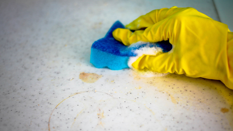 A person wearing yellow rubber gloves scrubbing at a yellow stain on a kitchen countertop