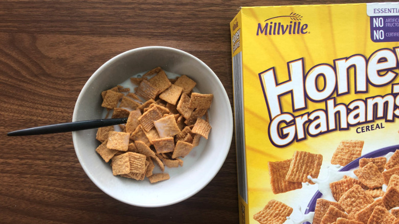 Honey Graham Squares cereal box beside a bowl with milk on a wood surface.