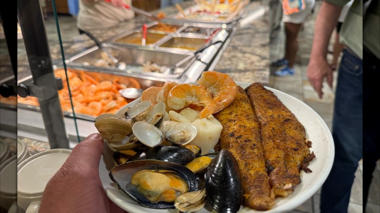 Hand holding plate of mussels, shrimp, and frie dfish in front of buffet line with seafood