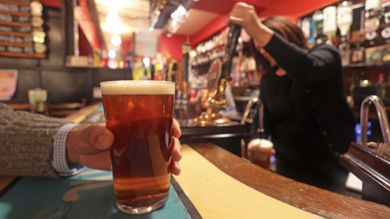 A hand holds a full pint of beer while a pub bartender pours another in the background