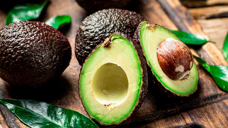 A ripe avocado that's cut in half exposing the pit on one side, sitting on a cutting board with two other avocados