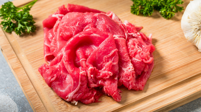 Pile of raw shaved beef on wooden board next to herbs and garlic