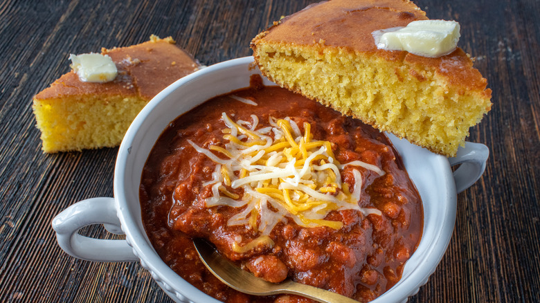 A white bowl of chili topped with cheese and served with two large pieces of cornbread