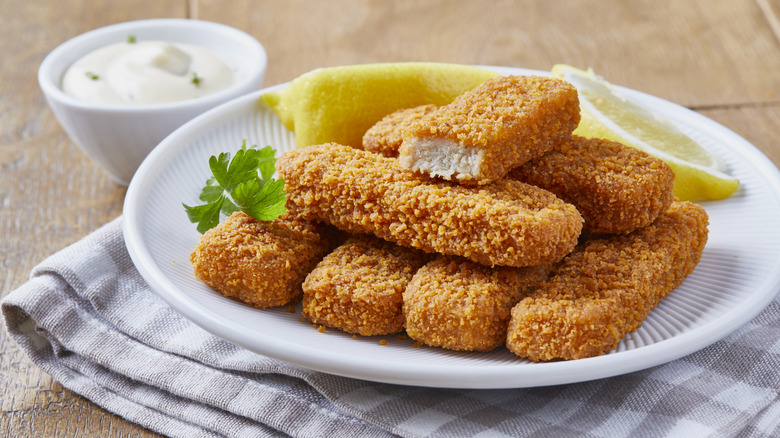 A white plate of fish sticks garnished with lemon and served with a small bowl of tartar sauce