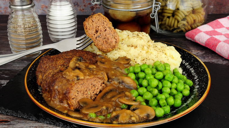 A plate of Salisbury steak, mashed potatoes, and green peas on a table next to salt and pepper shakers