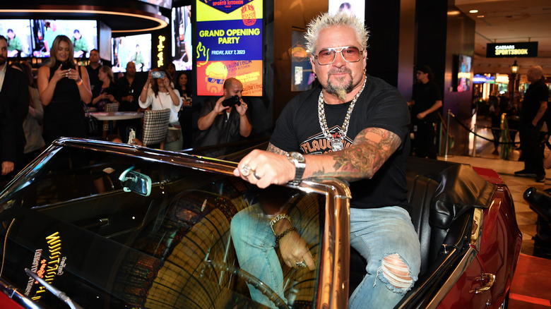 Guy Fieri sits in an open-topped car as the people surround him film him on their phones