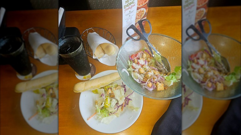 Meal at Olive Garden showing a bowl of salad with tongs, a breadstick basket, a soda, a menu, and a plate with salad and a breadsticks with mirror border