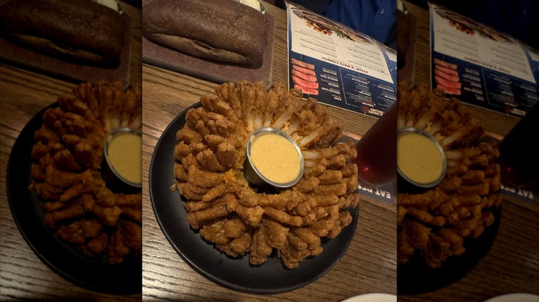 Wood dining table showing Outback Bloomin' Onion with dipping sauce, a loaf of brown bread, a drink, and a menu, with mirror border