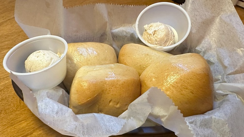 A basket of Texas Roadhouse rolls on parchment paper on a wood table with 2 sides of butter