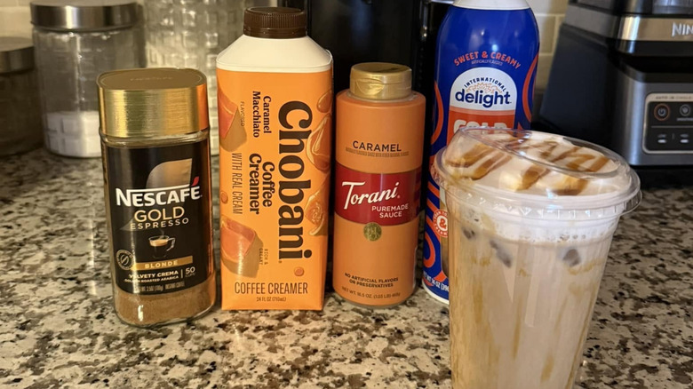 A carton of Chobani Caramel Macchiato Coffee Creamer on a counter next to an iced caramel macchiato, caramel syrup, whipped cream, and instant coffee
