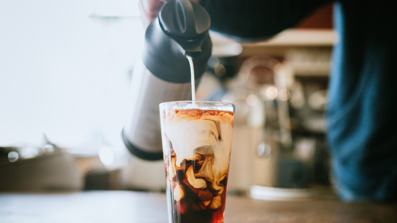 A close-up of a man pouring creamer into a tall glass of iced coffee