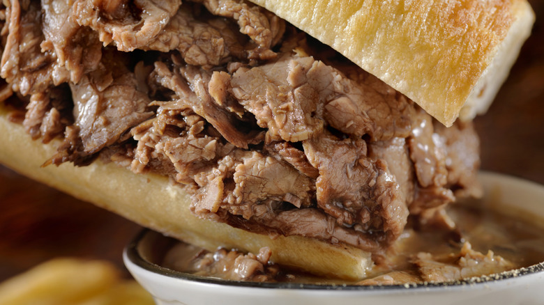 a beef dip sandwich, shown in a close-up image, with one corner dipped into the bowl of jus