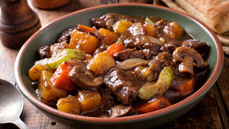 hearty beef stew in a terra-cotta bowl, shown on a rustic tabletop with a spoon, pepper grinder and the corner of a bread roll visible around it