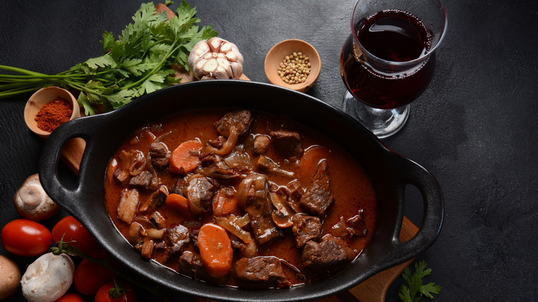 beef bourguignon in an oval dish on a black countertop, surrounded by ingredients including tomatoes, garlic, parsley, mushrooms, and a glass of wine