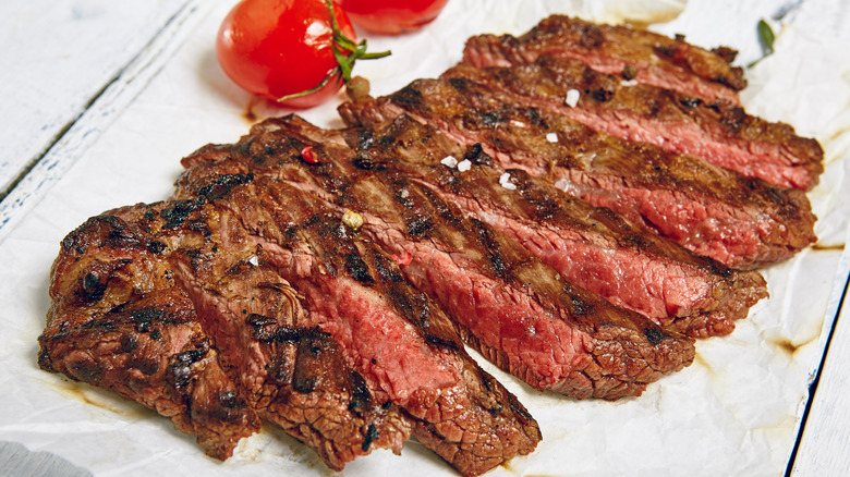 flank steak prepared as "London Broil," lightly charred and medium-rare, shown sliced on a white wooden rustic surface with cherry tomatoes visible behind