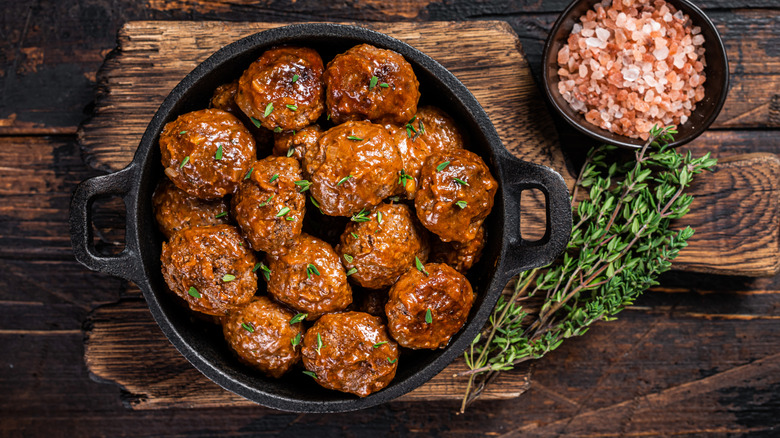 overhead view of meatballs in a cast-iron Dutch oven, on a rustic wooden surface, with a few sprigs of thyme and a bowl of coarse pink salt to its right