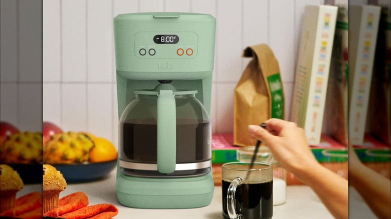 Green Bella 12 cup coffee maker on countertop with hand stirring coffee in a mug