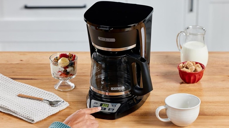 Hand turning on Black+Decker 12-cup programmable coffeemaker on tabletop with mug and breakfast items