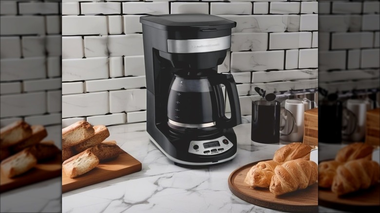 Black Hamilton Beach 12 cup programmable drip coffee maker on counter with mugs and pastries