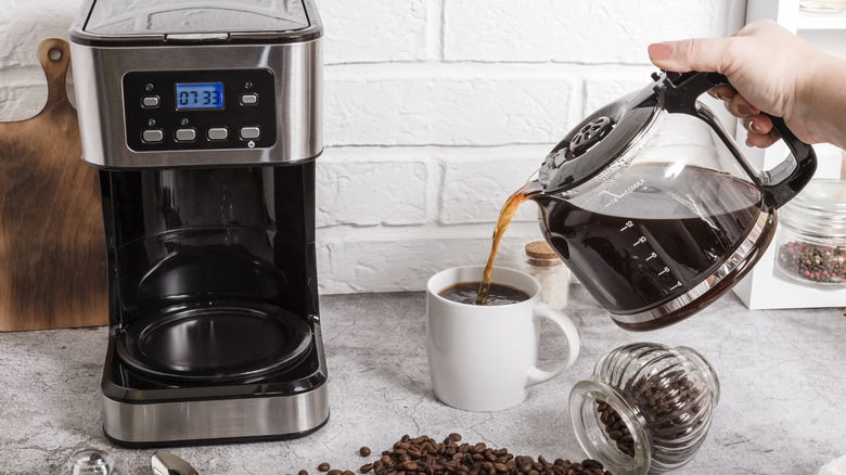 Hand pouring coffee from carafe into white coffee mug next to a drip coffee maker