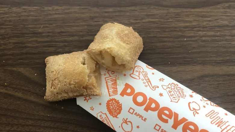 A Popeyes cinnamon apple pie propped up on the side of its orange and white container
