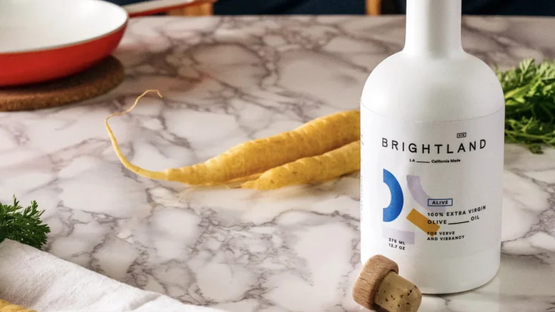 An uncorked bottle of Brightland Alive oil in front of some carrots on a marble counter