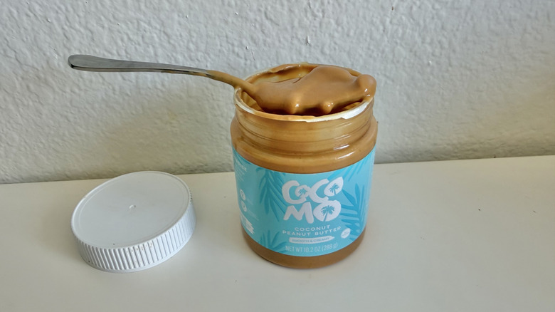 A spoonful of peanut butter resting on top of an open Cocomo Coconut Creamy Peanut Butter jar.