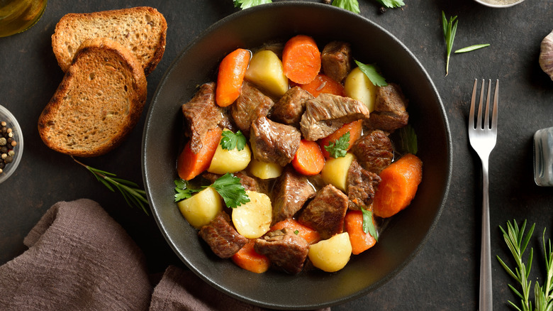 Bowl of pot roast next to fork, bread slices, and herbs