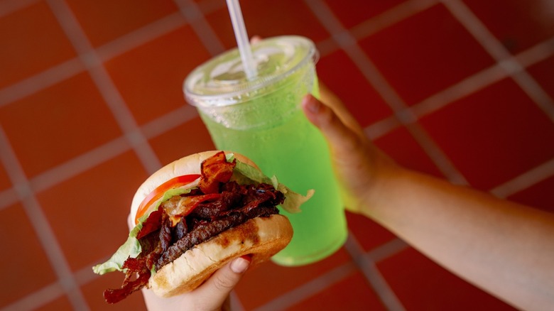 Hands holding a burger with bacon and tomato and a bright green drink