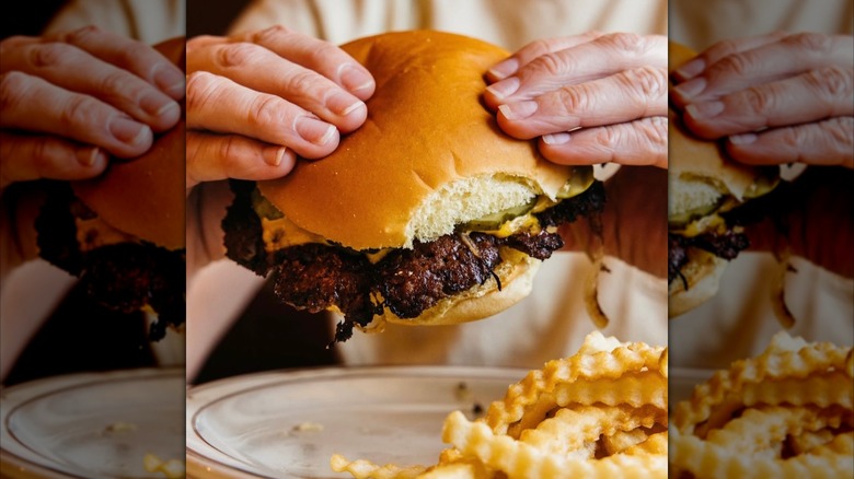 Hands holding a smashburger with fries on a plate