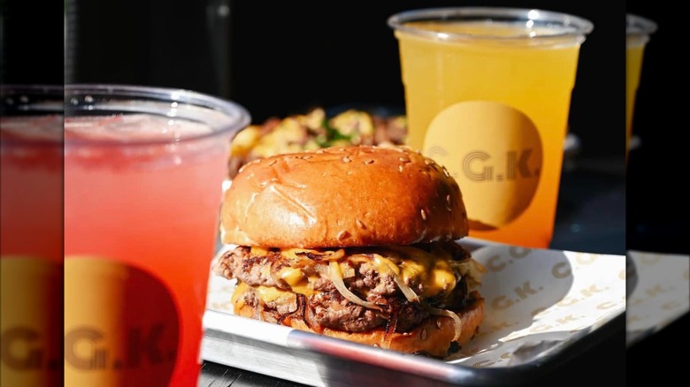 Burger on tray with paper and two plastic cups with orange and red drinks