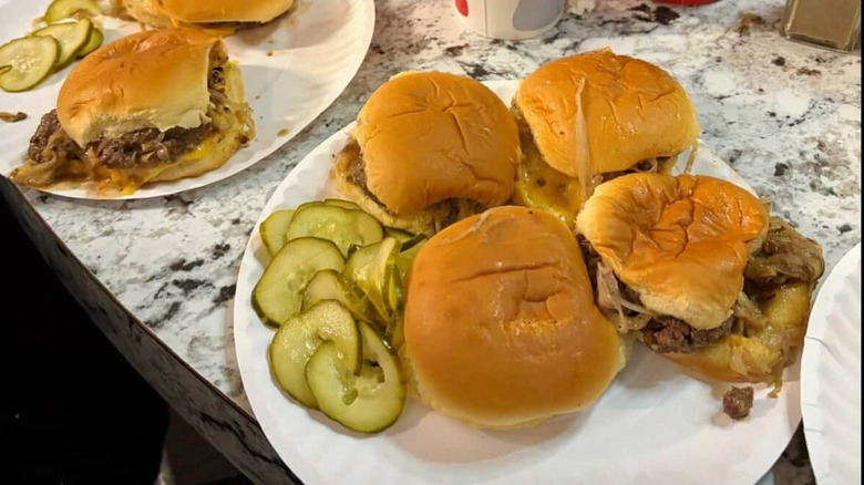 Paper plate of four hamburgers with pile of sliced pickles on the side